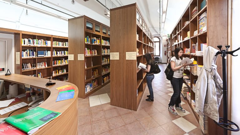 due giovani studentesse impegnate a consultare libri presi dagli ampi e alti scaffali della biblioteca dell'ateneo della Val d'Aosta